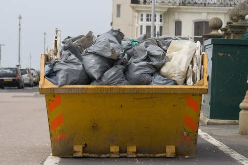A large yellow industrial skip filled with black and white rubbish bags is positioned on a paved street in a residential or commercial area near the coast, with buildings, parked cars, and a green waste collection bin visible in the background. The skip appears weathered with some dirt and scuff marks on its sides, and the bags are piled thickly, indicating a substantial disposal of waste. The surrounding environment includes a pavement with a curb, and the scene is taken in daylight, suggesting regular rubbish collection activity by commercial waste services in Brompton. The overall setting reflects typical urban waste management practices relevant to local rubbish removal services like Commercial Waste Brompton.