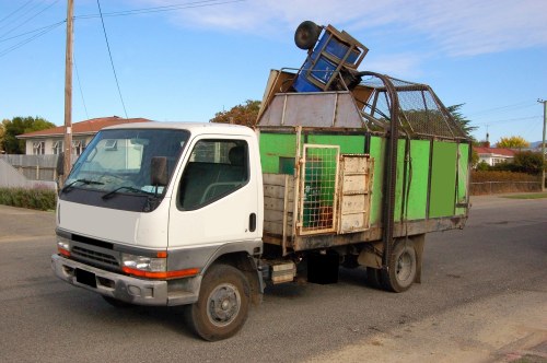 A small commercial flatbed truck parked on a residential street, with a white cab and a green cargo area constructed from metal panels and wooden slats. The cargo bed is loaded with various discarded items, including a metal chair, a plastic pallet, and other miscellaneous waste materials. The truck is positioned in an area with a paved surface, and in the background, there are houses with tiled roofs, trees, and utility poles under a partly cloudy sky. The vehicle appears used, with some rust visible on the metal parts, and the load is stacked haphazardly. The scene suggests a rubbish removal operation, typical of services provided by Commercial Waste Brompton in the local area, possibly near Brompton or a nearby postcode district. The environment is quiet, with no other vehicles or people immediately visible, emphasizing the purpose of waste collection and clearance.