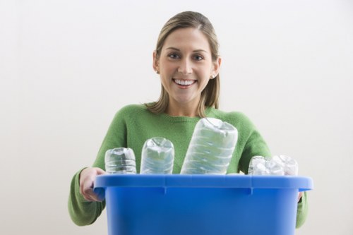 A young woman with light brown hair, smiling and wearing a bright green sweater, holds a blue recycling bin filled with empty, transparent plastic bottles of various sizes and shapes. The bottles, made from clear PET plastic, display slight textural details such as ridges and smooth surfaces, and are positioned upright in the bin, some leaning against each other. The background is plain white, highlighting the focus on the woman and the bin. The setting appears to be indoors, possibly in a home or office environment, with good lighting illuminating her face and the contents of the bin. This image emphasizes the importance of plastic bottle recycling, aligning with rubbish removal services offered by Commercial Waste Brompton in the Brompton area, subtly supporting local waste management efforts while depicting environmentally conscious disposal practices.