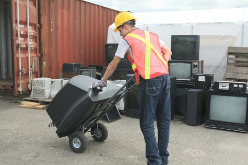 A person wearing a yellow safety helmet and a high-visibility vest over a white T-shirt is seen pushing a black wheeled rubbish bin, which is tilted forward as they move it across an outdoor area. Behind them, there are several old electronic appliances, such as CRT televisions and audio equipment, stacked against a metal shipping container and a wooden pallet. The environment appears to be a commercial or industrial yard, possibly part of waste disposal or disposal site near Brompton, with the ground surface made of asphalt or concrete. The scene is lit with natural daylight, and the overall setting suggests a rubbish removal process as conducted by Commercial Waste Brompton, specifically handling electronic waste and general rubbish disposal.