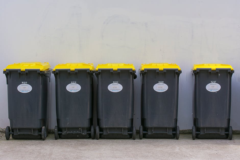 A small, old blue flatbed truck parked on the side of a street, loaded with a large amount of mixed rubbish including cardboard boxes, black garbage bags, and other waste materials. The truck's side p