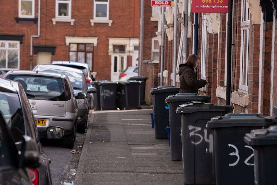 An overflowing outdoor waste collection point featuring a large metal mixed paper and cardboard bin with its lid open, revealing scattered paper waste and packaging inside. Surrounding on the pavement