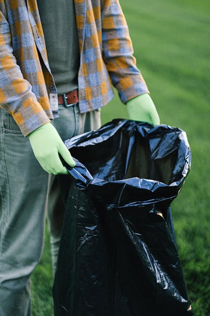 A worker wearing high-visibility clothing is operating a large, red waste collection vehicle parked on the side of a street. The rear of the vehicle features an open compactor section where the worker